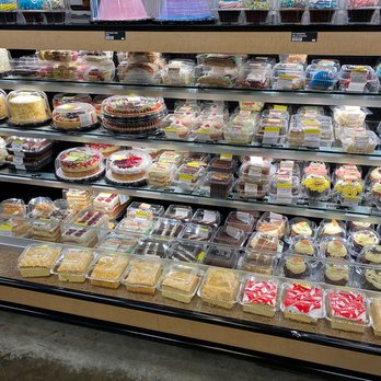 Mariano's - Bakery section in a store showing multiple shelves filled with pre-packaged cakes, slices, and cupcakes. The top shelf has decorated cupcakes under clear plastic covers, while the lower shelves contain slices of cake and whole cakes in plastic containers. The display is organized with different types of desserts, including fruit-topped cakes and frosted cupcakes, in a retail environment with a tiled wall background. - Grocery Near Me - Westchester, Illinois