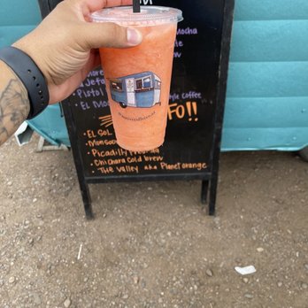 Native Coffee - The Valley drink. It's a pomegranate/mango drink with chamoy. It's nice, refreshing and yummy - A person holds a colorful smoothie in a clear cup with a black straw, featuring a caravan logo, in front of a blackboard menu at Native Coffee. - Coffee & Tea Near Me - Tolleson, Arizona