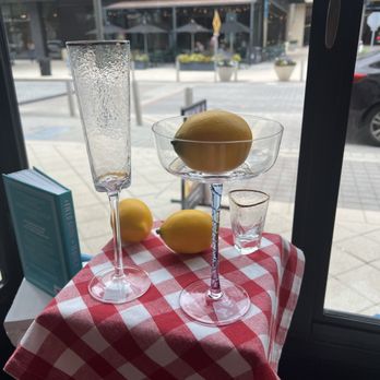 Read Between the Lines - Lemons and glassware, including a tall champagne flute, a coupe glass with a lemon, and a small shot glass, are arranged on a red and white checkered tablecloth inside a restaurant. The table is near large windows, showing a street scene with a Shake Shack restaurant across the street, outdoor seating, and parked cars. The setup suggests a decorative or promotional display. - Cards & Stationery Near Me - Plano, Texas