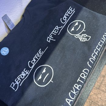 Blackbird Coffee House - Hehe, so cute - A black T-shirt features white text and illustrations, including a smiley face and coffee-related phrases, laid on a surface. - Coffee & Tea Near Me - Albuquerque, New Mexico