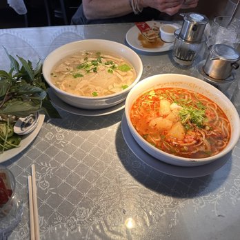Cafe Da Lat - Chicken noodle soup (left) Spicy beef stew (right) Ice coffee - Two bowls of Vietnamese noodle soup, one with clear broth and the other with spicy red broth, are on a table with fresh herbs, condiments, and glasses of ice water. - Vietnamese Near Me - Albuquerque, New Mexico