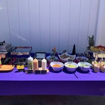Chuy Tacos - The whole shabang - An outdoor buffet table displays bowls of food, condiments, and small cups, with a purple tablecloth and a white wall in the background. - Caterers Near Me