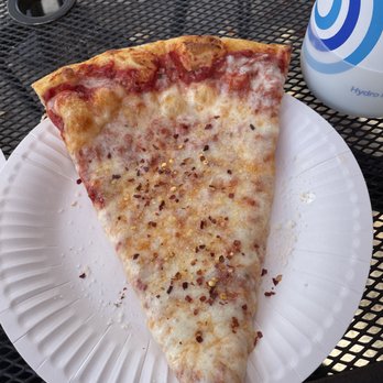 Sy's New York Pizza - A single large slice of cheese pizza with a golden crust, melted cheese, and red pepper flakes is served on a white paper plate. The pizza is on a black metal outdoor table, with a blue and white water bottle and a black circular hole in the table visible nearby. - Pizza Near Me - Eugene, Oregon