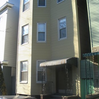 MYG Carpentry Service - Before pic of Chelsea St - A multi-story residential building features yellow horizontal siding, with several windows on each floor. The entrance has a small set of stairs leading up to a door, shaded by a striped awning. The building is located in a neighborhood with other similar structures, and a parked car is visible in the foreground. The image captures the building's exterior and part of the surrounding area. - Carpenters Near Me