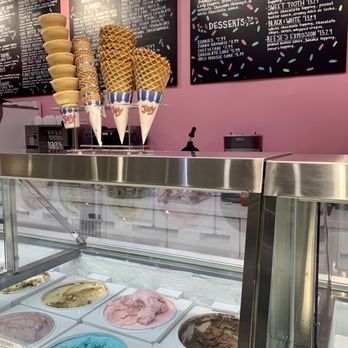The Creamery - Ice cream cones and waffle cones are arranged on top of a stainless steel gelato display case at an ice cream shop. The cones are in paper holders with the brand name 'Joy.' Behind the cones, there are menu boards listing flavors and desserts, with a pink wall as the background. The gelato case contains various flavors of ice cream, including pink, blue, and brown varieties, visible through the glass. The setting appears to be a modern ice cream shop near Brandeis University. - Ice Cream & Frozen Yogurt Near Me - Waltham, Massachusetts