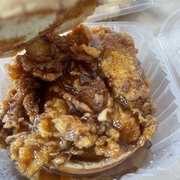 Crazy N Luv Sandwich - The image depicts a chicken sandwich with crispy, seasoned fried chicken pieces topped with gravy inside a clear plastic container. The sandwich is partially covered by a bun at the top, and the container is placed on a light-colored surface. The focus is on the fried chicken and gravy, highlighting the texture and seasoning.