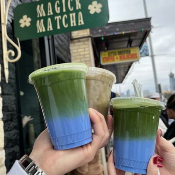 Magick Matcha - Magick matcha and hojacha latte - Three matcha drinks with layered colors are held by two people outside Magick Matcha, a shop in Austin, TX, with a sign and storefront visible. - Coffee & Tea Near Me - Austin, Texas
