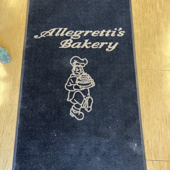 Allegretti's Bakery - Entry way - A rectangular black doormat features the Allegretti's Bakery logo and name in white script, with a cartoon baker holding a cake. - Bakeries Near Me - Norridge, Illinois