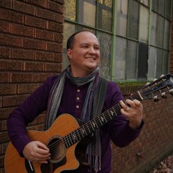 Eli Conley - Image Description: Eli, a white man, is smiling and playing acoustic guitar in front of a run down building.
