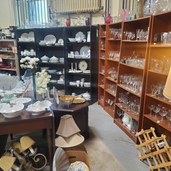 Serendipity Shoppe - The image shows the interior of a thrift store with multiple shelves filled with glassware, dishes, and decorative items. A table in the foreground holds vases, bowls, and a flower arrangement. The floor has a box containing lampshades and other miscellaneous objects. The store has a window with bars, a radiator, and a mix of furniture and household items arranged for sale. - Thrift Stores Near Me - Buffalo, New York