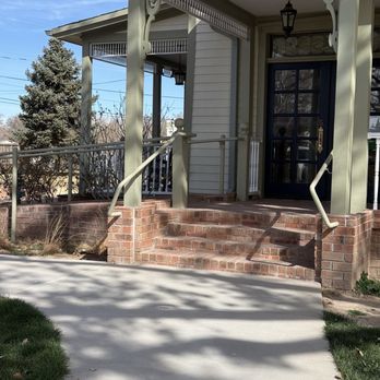 Heritage House - Outdoor entrance - A house entrance features brick stairs leading up to a porch with decorative trim and a black door with glass panels. A hanging lantern light is above the door, and the porch has a railing with vertical balusters. The house has light-colored siding, and there are trees and power lines visible in the background. Shadows from nearby trees cast patterns on the sidewalk and grass. - Venues & Event Spaces Near Me - Corrales, New Mexico