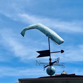 Whaling Museum - Weathervane a top the roof - A whale-shaped weather vane is installed on the roof of a building, with directional indicators (N, E, S, W) and a globe-like base. The weather vane is set against a clear blue sky with some wispy clouds, and the roof has visible gutters and a small vent. - Museums Near Me - Nantucket, Massachusetts