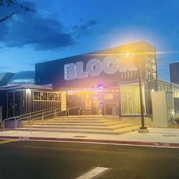 The Block building has a large sign spelling 'BLOCK' on its facade, illuminated by exterior lighting. The building's entrance is accessed via a set of stairs with handrails, and a streetlamp is positioned nearby. The sky is partly cloudy with a blue hue, indicating early evening or dusk. The parking lot in front is mostly empty, with visible parking lines and a curb marked 'FIRE LANE.'