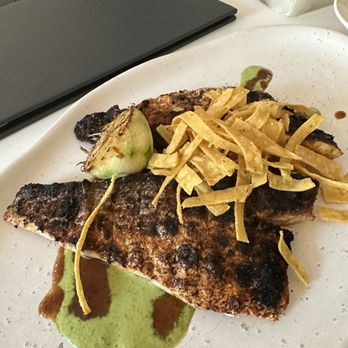 SARA - Sea bass - The image shows a white plate with a grilled fish fillet that has a dark, crispy exterior. It is garnished with fried plantains on top and a piece of green fruit, possibly a plantain or avocado. There is a green sauce with brown drizzles underneath the fish, and the plate is set on a white table with a black menu or folder nearby. The background includes chairs and part of a restaurant interior. - Canadian (New) Near Me - Toronto, Ontario