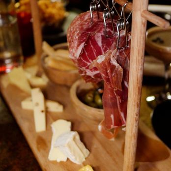 CODE RED - Cheese and Charcuterie - A wooden charcuterie board displays slices of cured meats hanging from a wooden and metal rack, with cheese slices, mustard, and butter placed on the board. The background includes blurred drinks and food items, suggesting a dining or bar setting. - Cocktail Bars Near Me - Washington, DC, District of Columbia