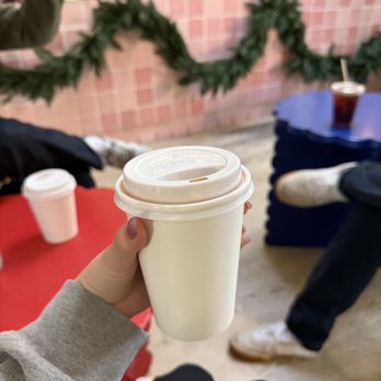 Loveless Coffees - A person is holding a white disposable coffee cup with a lid in a coffee shop with pink tiled walls and green garland decorations. - Coffee Roasteries Near Me - Brooklyn, New York