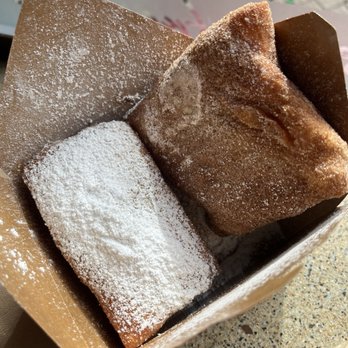 Powderface - Beignets (2 powder sugar traditional and 1 cinnamon sugar) - Two beignets, one rectangular and one square, dusted with powdered sugar, are placed in a paper bag on a speckled surface. - Coffee & Tea Near Me - Oakland, California