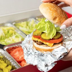A close-up photo of a person putting the top bun of their cheeseburger in front of a Five Guys catering box of toppings.