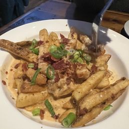 Loaded fries . Loaded with flavor - A white round plate contains loaded fries topped with melted cheese, crispy bacon bits, and chopped green onions. A fork is inserted into the dish, and the fries are coated in a creamy sauce. The setting appears to be a restaurant with a wooden table and dim lighting.