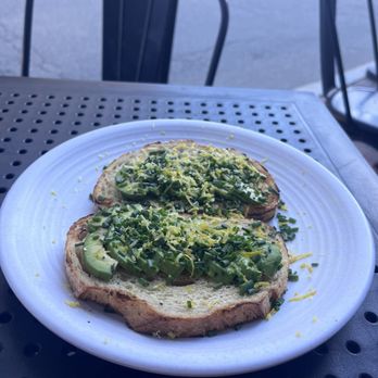 Vester - Two slices of toasted bread are topped with sliced avocado, chopped green onions, and lemon zest. The plate is placed on a perforated metal table outside, with chairs and a street visible in the background. The setting appears to be an outdoor dining area, possibly at a cafe or restaurant. - Cafes Near Me - Cambridge, Massachusetts