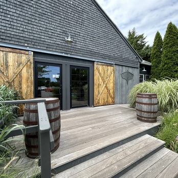 Nomans - Entrance - An outdoor wooden deck features two wooden barrels, lush plants, and a gray building with sliding glass doors and wooden barn-style doors. - American Near Me - Oak Bluffs, Massachusetts