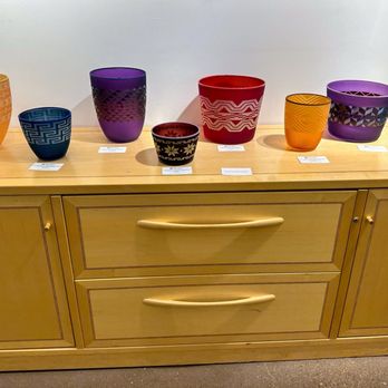 Blue Rain Gallery - Multiple glass bowls in different colors, including orange, blue, purple, red, and yellow, are displayed on a wooden cabinet in an art gallery setting. Each bowl has unique patterns and is accompanied by small informational labels. The display is part of the gallery's collection of contemporary Native American art and studio glass. - Art Galleries Near Me - Santa Fe, New Mexico