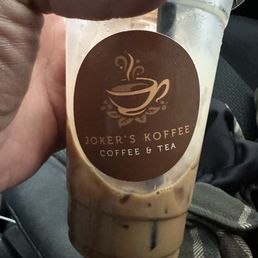 Vietnamese Coffee. - A clear plastic cup containing iced coffee with milk, featuring a Joker's Koffee logo and label, held by a person's hand.