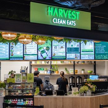 Harvest Clean Eats - Storefront - The image shows the exterior of Harvest Clean Eats, with a prominent green sign displaying the business name. Below the sign, digital menu boards are visible, showcasing various food and drink options. The interior counter area is partially visible, with staff members preparing orders. The decor includes hanging lights and greenery, creating a modern and fresh appearance. - Juice Bars & Smoothies Near Me - Toronto, Ontario