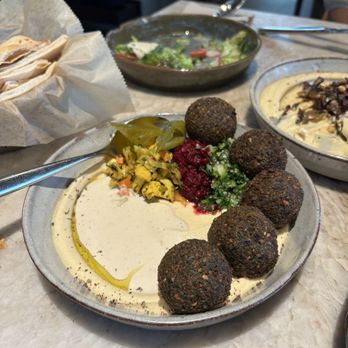 Rumis Kitchen - Falafel - A dish featuring falafel balls served with pickles, beet hummus, and chopped vegetables, on a restaurant table with other dishes and utensils. - Persian/Iranian Near Me - Houston, Texas