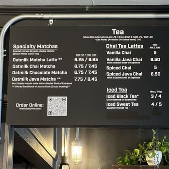 Two Cities Coffee Roasters - Matcha & Tea Menu - The image shows a black menu board hanging above a coffee shop counter, listing tea, matcha, and latte options with prices. The menu includes specialty matchas, chai tea lattes, and iced teas, with details about sizes and ingredients. A QR code for online ordering is also visible. The background features a ceiling light, some plants, and part of the coffee shop interior. - Coffee Roasteries Near Me - Clovis, California