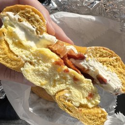 Southern Tradition with Bacon on a Jalapeño Cheddar Bagel + cream cheese added - A bagel sandwich with scrambled eggs, crispy bacon, and cream cheese is held in a person's hand. The bagel is sliced in half, showing the filling inside. The background includes foil wrapping and part of a person's lap, indicating the sandwich is being eaten in a casual setting.