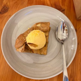 Sable Kitchen + Bar - Apple pie with vanilla ice cream on top, served on a gray ceramic plate, with a metal spoon on the side, placed on a wooden table. - Salad Near Me - Lawrence, Kansas