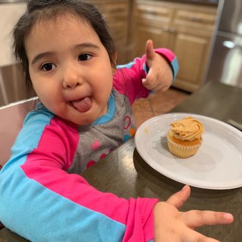Brooklyn Cupcake - A young girl with dark hair and light skin is seated at a table in a kitchen or dining area. She is wearing a long-sleeve shirt with pink, blue, and gray colors. She is making a playful face by sticking out her tongue and giving a thumbs-up with her right hand. In front of her, on a white plate, there is a cupcake with light brown frosting. The background shows wooden cabinets and kitchen appliances, indicating an indoor setting. - Cupcakes Near Me - Brooklyn, New York