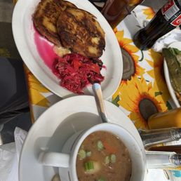 Green Chile Beef Stew - The image shows a plate with two Salvadoran pupusas served with pickled cabbage, a bowl of green chile beef stew garnished with chopped green onions, and two beverages—iced tea with lemon and a bottle of soda—on a table with a sunflower-themed tablecloth. The setting appears to be outdoors, with sunlight illuminating the food and drinks.