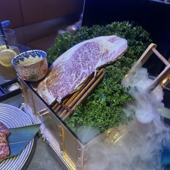 Y Space BBQ - A piece of Wagyu beef with extensive marbling is placed on a bed of curly kale leaves inside a decorative metal box. The box emits fog from dry ice, creating a smoky effect. The setting appears to be a restaurant, with a person in the background and a glass of beverage nearby. The presentation emphasizes the quality and visual appeal of the beef. - Barbeque Near Me - Flushing, New York