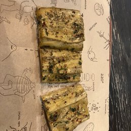 Spicy Eggplant - Three slices of eggplant, seasoned with herbs, chili flakes, and oil, are arranged on paper featuring hand-drawn illustrations of chefs, food items, and kitchen tools. The paper is on a dark wooden surface, and the slices appear grilled or roasted with visible seasoning and herbs.