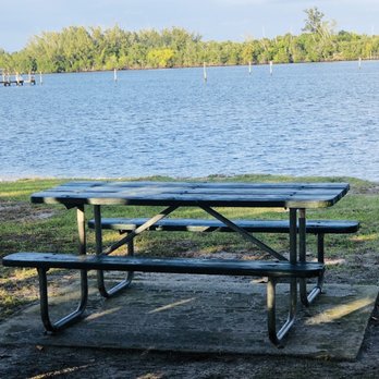 Cove Road Park - A weathered wooden picnic table with attached benches is placed on a concrete pad beside a lake, with trees and docks visible in the background. - Parks Near Me - Stuart, Florida