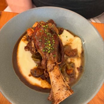 Sable Kitchen + Bar - A lamb shank with sauce is served on a plate with mashed potatoes and vegetables, garnished with chopped green onions. - Salad Near Me - Lawrence, Kansas