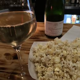 A glass of white wine, a bottle of sparkling wine, and a bowl of popcorn are placed on a wooden table in a dimly lit setting.