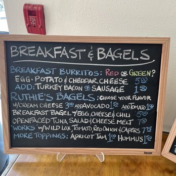 Ruthie's Bagels - Breakfast menu - A chalkboard menu at Ruthie's Bagels features handwritten items including breakfast burritos, bagels, and toppings. The menu lists options such as egg, potato, cheddar cheese, turkey bacon, sausage, and various bagel flavors with prices. The board is framed in wood and positioned on a table, with a window and a fire alarm visible in the background. - Bagels Near Me - Albuquerque, New Mexico