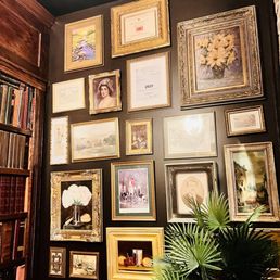 The image shows a black wall adorned with multiple framed artworks, including paintings, photographs, and certificates, arranged in a gallery style. To the left, part of a wooden bookshelf filled with books is visible. A green plant with large, fan-shaped leaves is positioned at the bottom right corner, partially obscuring some frames. The setting appears to be an interior space with warm, subdued lighting.
