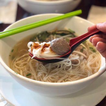 Saigon Cafe - Saigon House Special - A bowl of Vietnamese pho with clear broth, rice noodles, herbs, and a meatball on a spoon held by a hand. Chopsticks are resting on the bowl's edge, and another bowl of pho is visible in the background. The setting appears to be a restaurant table. - Vietnamese Near Me - Eugene, Oregon
