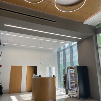Rush South Loop - The image shows a modern indoor space with a high ceiling and large glass windows allowing natural light. There are two wooden doors on the back wall, a curved wooden reception desk in the center, and vending machines to the right. The ceiling has contemporary circular and linear light fixtures, and the floor is made of polished material. The space appears to be a lobby or waiting area, possibly in an office or public building. - Doctors Near Me - Chicago, Illinois