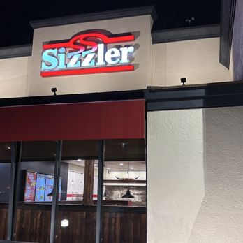 Sizzler - Nighttime view of a Sizzler restaurant's exterior, featuring a lit sign and large glass windows showing part of the interior. - Seafood Near Me - Merced, California