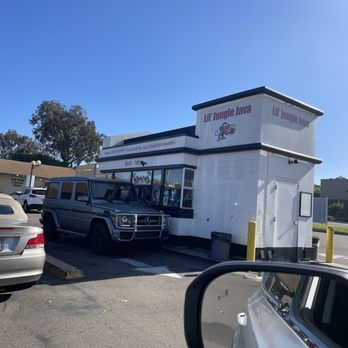 Lil' Jungle Java -  car parked in front of a restaurant - A small drive-through coffee stand named Lil' Jungle Java features a white exterior with black trim and a cartoon monkey logo. The stand is located in a parking lot with several parked cars, including a gray Mercedes-Benz G-Class SUV. The sky is clear and blue, indicating a sunny day. The stand offers beverages and snacks, with signage indicating options like coffee ice cubes and smoothies. The image captures the storefront and surrounding vehicles. - Coffee & Tea Near Me - Encinitas, California