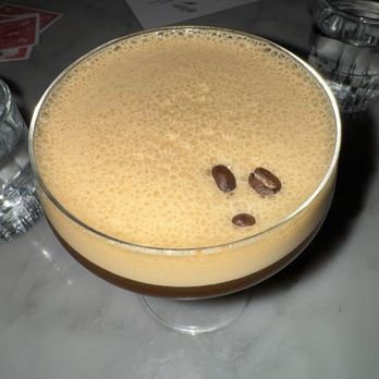 Root & Flower - Espresso martini - A coffee drink with a frothy, light-colored top layer and a few coffee beans placed on the surface as garnish. The drink is served in a clear glass on a table, with water glasses and a menu visible in the background. The setting appears to be a café or restaurant environment. - Wine Bars Near Me - Vail, Colorado