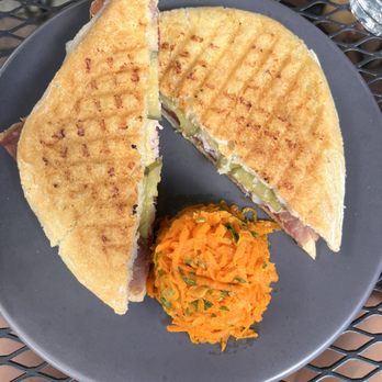 Tilman's - Yummmm- the perfect proportion for lunch - A grilled sandwich with visible layers of meat and pickles, cut in half, served with a side of shredded carrot salad on a dark plate. - Wine Bars Near Me - Charlottesville, Virginia