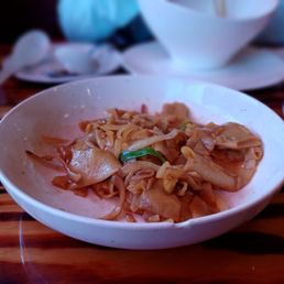 Noodles - A bowl of Asian-style noodles with sauce and vegetables is placed on a wooden table, with other dishes and utensils visible in the background.