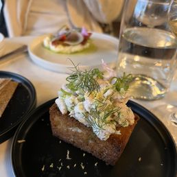 Shrimp Toast - A rectangular piece of toasted bread with a golden-brown crust is topped with a creamy salad mixture, garnished with fresh herbs such as dill. The dish is presented on a black plate, with a glass of water and other dishes visible in the background, indicating a dining setting.
