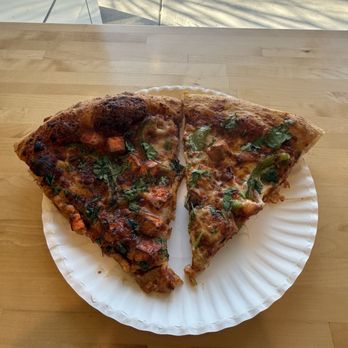 Desi Boys Pizza - Paneer and butter chicken - Two slices of pizza with toppings, including green herbs, are arranged on a white paper plate on a wooden table. The pizza has a slightly charred crust and appears to be freshly baked. The background shows a window with sunlight and shadows cast on the surface outside. - Pizza Near Me - Chicago, Illinois